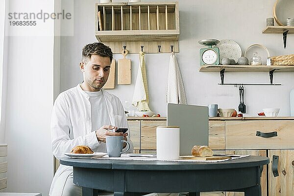 Junger Mann mit Smartphone sitzt in der Nähe von Tisch mit Frühstück Laptop Küche