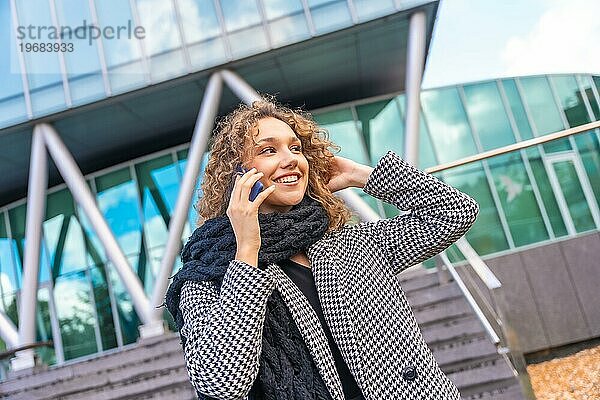 Glückliche junge Geschäftsfrau in eleganter warmer Kleidung  die während des Herbstes in der Stadt telefoniert