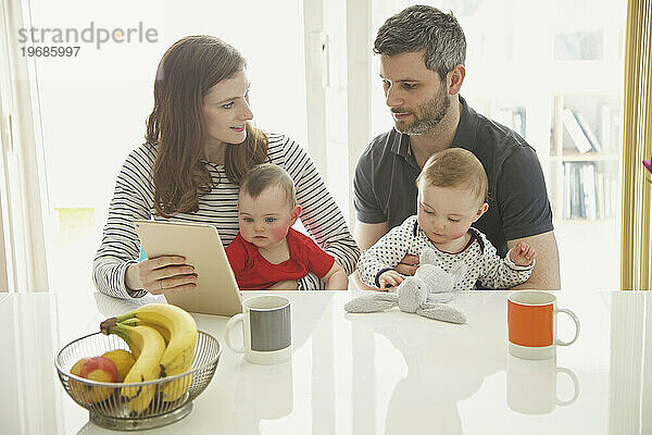 Paar sitzt mit Zwillingsbabys am Tisch und nutzt digitales Tablet