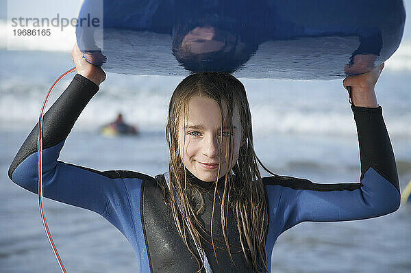 Lächelndes und nasses Mädchen  das ein Surfbrett auf dem Kopf hält und aus dem Meer kommt