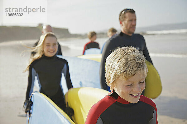 Junge und Mädchen tragen Surfbretter am Strand  gefolgt von Menschen