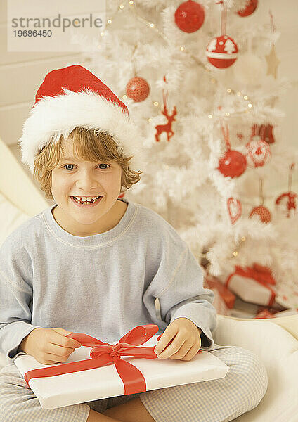 Ein Junge mit rot-weißer Pelzmütze sitzt am Weihnachtsbaum und packt ein Geschenk aus