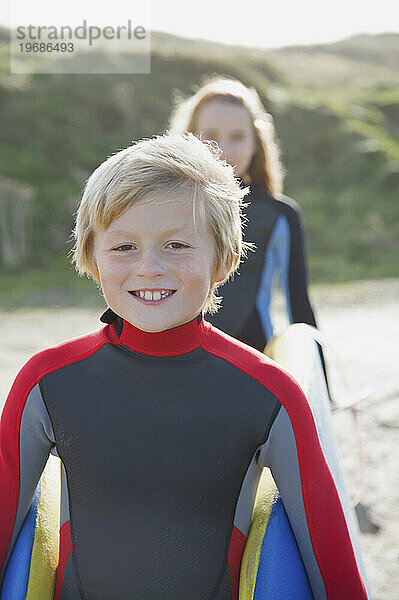 Lächelnder Junge im Neoprenanzug  der ein Surfbrett mit einem Mädchen trägt