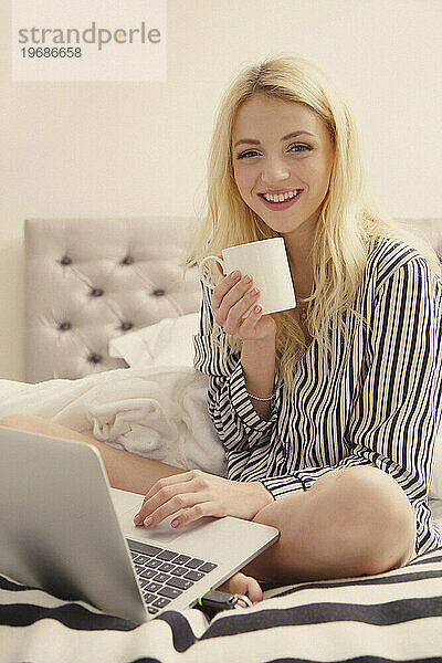 Lächelnde junge Frau sitzt mit Laptop im Bett
