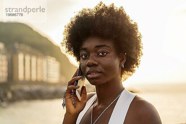 Nahaufnahme einer ernsten und schönen afrikanischen jungen Frau  die bei Sonnenuntergang im Freien mit dem Handy spricht