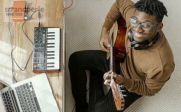 Draufsicht auf einen lächelnden männlichen Musiker  der zu Hause Gitarre spielt und mit einem Laptop mischt