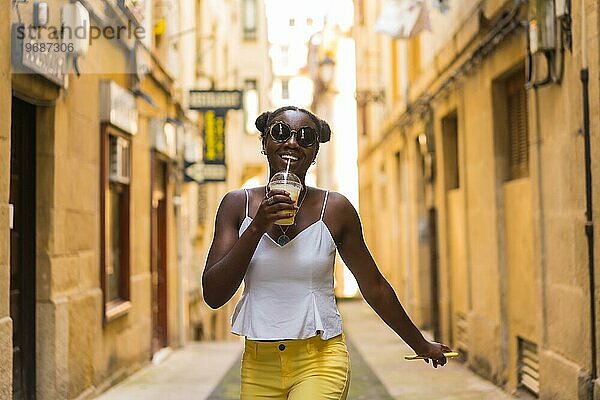 Coole afrikanische junge Frau trinkt Saft  während sie fröhlich auf einer Straße spazieren geht