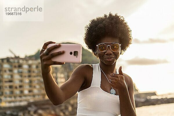 Afrikanische Frau gestikuliert  dass es in Ordnung ist  ein Selfie im Freien neben dem Meer bei Sonnenuntergang zu machen