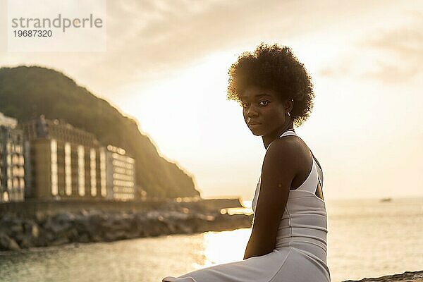 Afro Schönheit Frau drehen  um in die Kamera schauen  während der Blick auf den Ozean bei Sonnenuntergang