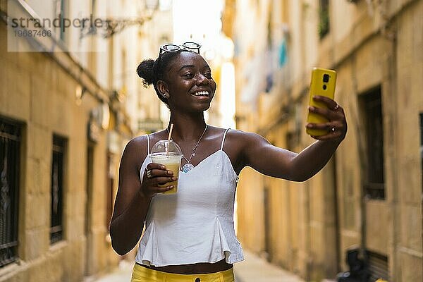 Lächelnde junge afroamerikanische Frau  die ein Selfie mit einem leckeren Milchshake auf der Straße in der Stadt macht