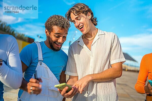 Lächelnde und abgelenkte multiethnische junge Männer  die im Freien neben ihren Freunden telefonieren