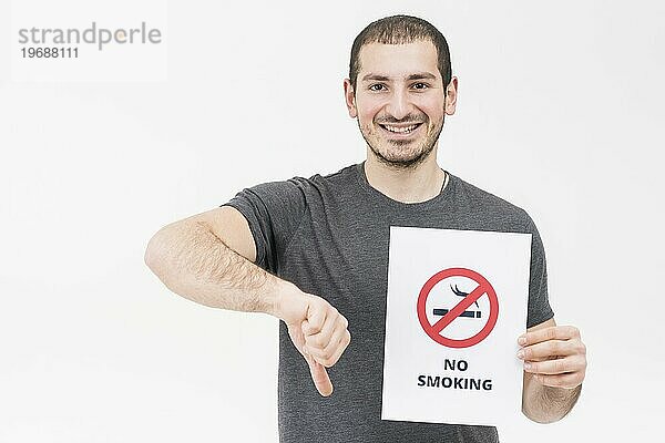 Glücklicher junger Mann  der ein Nichtraucherzeichen hält und den Daumen nach unten zeigt  isoliert vor einem weißen Hintergrund