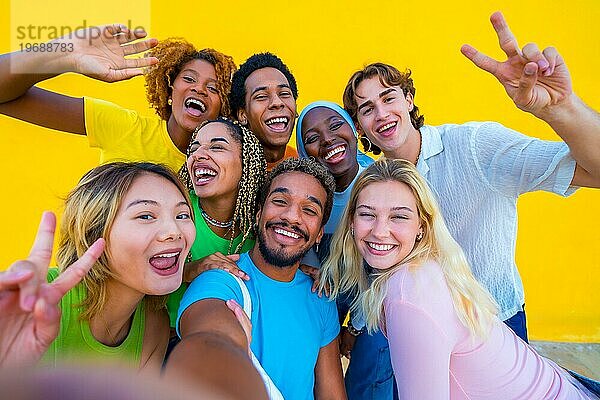 Multiethnische Freunde machen ein Selfie neben einer gelben Wand und gestikulieren dabei Frieden