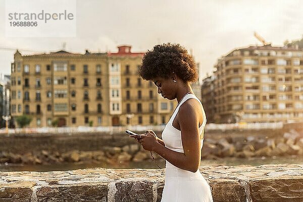 Seitenansicht einer stilvollen afro Frau mit Telefon zu Fuß im Freien während des Sonnenuntergangs neben dem Meer