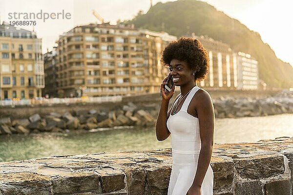 Glückliche afrikanische Frau in schäbigem Sommerkleid  die bei Sonnenuntergang im Freien spazieren geht und sich mit dem Handy unterhält