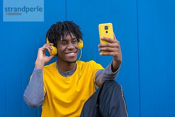 Glücklicher Afro Mann  der ein Selfie macht  während er auf dem Boden sitzend vor einem blauen Hintergrund Musik hört