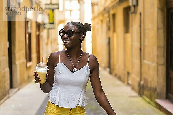 Glückliche afrikanische junge Frau  die einen eisgekühlten Milchshake trinkt  während sie eine Straße entlanggeht