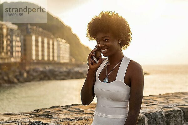 Horizontales Foto mit Kopierbereich einer lächelnden Afro Frau  die bei Sonnenuntergang im Meer mit dem Handy spricht