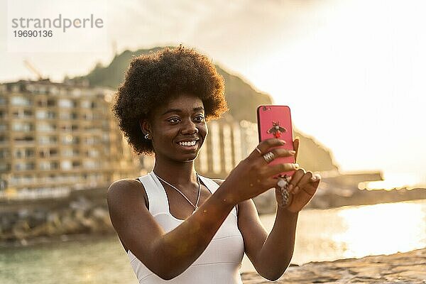 Glückliche Afro Frau im weißen Kleid macht ein Selfie bei Sonnenuntergang am Meer