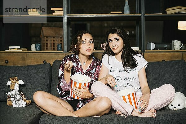 Zwei verängstigte Frauen sitzen mit Popcorn auf dem Sofa und sehen fern