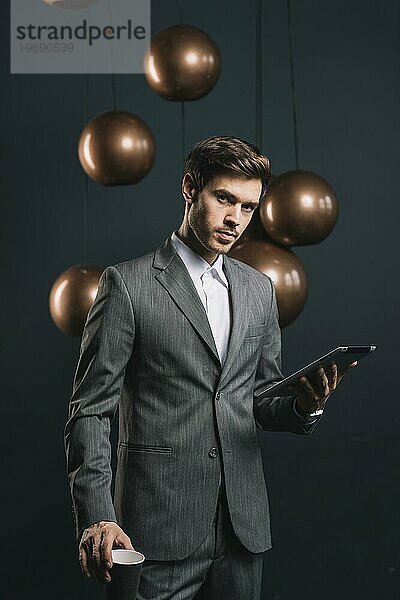 Selbstbewusster junger Mann mit Einwegbecher und digitalem Tablet vor dunklem Hintergrund