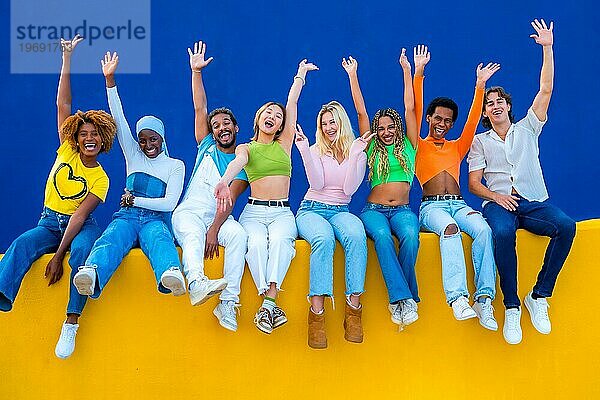 Junge multiethnische Freunde  die sich die Hände reichen und lachend auf einer bunten Wand sitzen