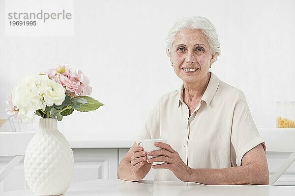 Ältere Frau hält Tasse Kaffee mit Blumenvase weißen Tisch