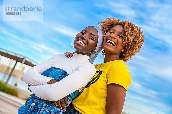 Afrikanische Freundinnen stehen zusammen und lächeln in die Kamera in einem städtischen Raum mit blauem Himmel