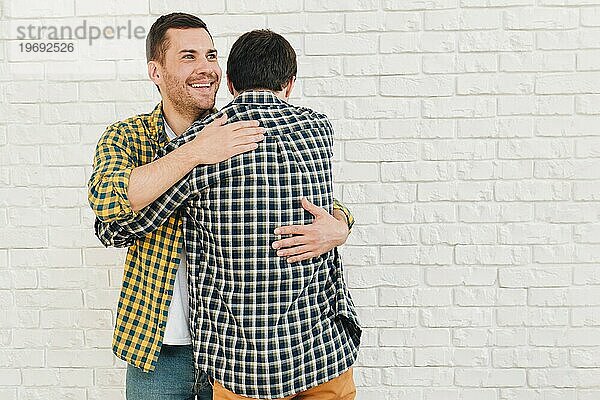 Lächelnde Porträt Mann gibt umarmen seinen Freund gegen weiße Backsteinmauer