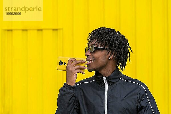 Afrikanischer junger Mann  der eine Sprachnachricht mit dem Handy neben einer gelben Stadtmauer verschickt