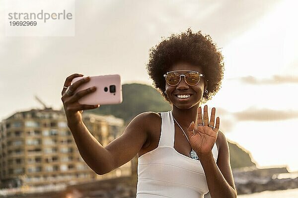 Porträt einer stilvollen afrikanischen Frau  die während eines Videogesprächs am Meer winkt