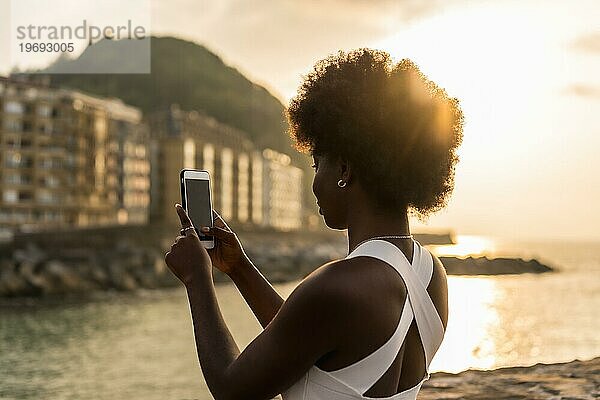 Rückansicht einer afrikanischen stilvollen jungen Frau beim Fotografieren des Sonnenuntergangs am Meer