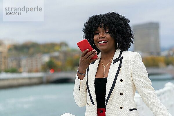 Elegante afroamerikanische Geschäftsfrau  die eine Sprachnachricht im Freien neben einem städtischen Fluss sendet