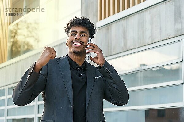 Horizontales Porträt eines glücklichen arabischen jungen Geschäftsmannes  der erfolgreich gestikuliert  während er ein Telefon benutzt