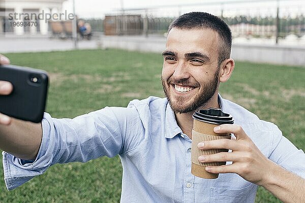 Lächelnder Mann  der ein Selfie macht  während er einen Einweg Kaffeebecher im Park hält