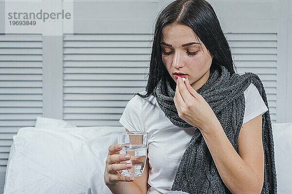 Frau mit Erkältung  die ein Glas Wasser in der Hand hält und Medizin einnimmt
