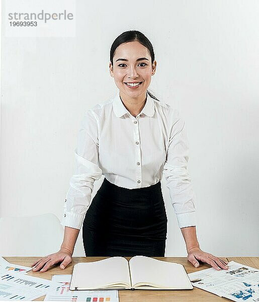 Lächelndes Porträt junge Geschäftsfrau stehenden Schreibtisch gegen weiße Wand