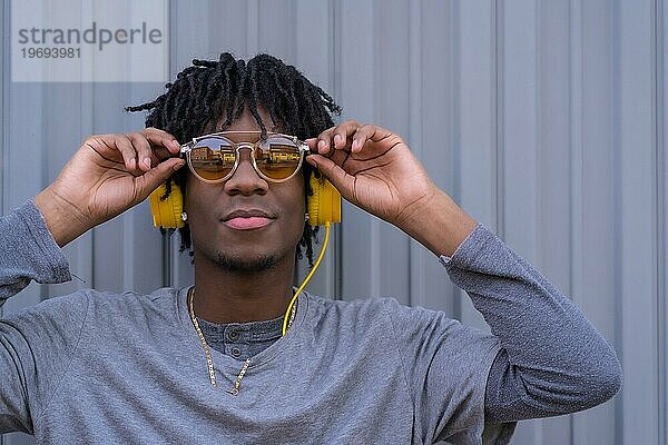 Selbstbewusster afrikanischer junger Mann mit Sonnenbrille und Kopfhörern vor einem grauen städtischen Hintergrund