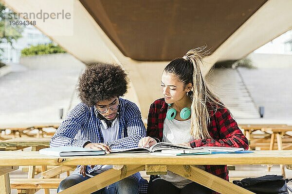 Verschiedenes junges Paar sitzt auf einer Holzbank und studiert zusammen auf dem Universitätscampus