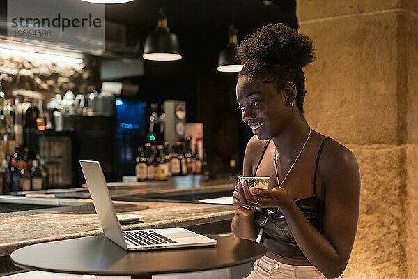 Seitenansicht einer glücklichen und jungen entspannten Frau  die während eines Online Meetings an einer Bar sitzt