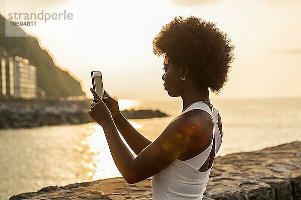 Rückansicht einer afrikanischen Frau  die den Sonnenuntergang im Meer fotografiert