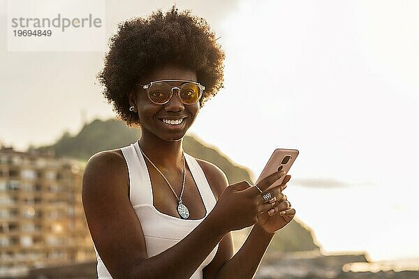 Stilvolle afrikanische Frau lächelt in die Kamera  während sie bei Sonnenuntergang im Freien telefoniert