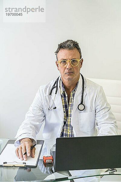 Männlicher Arzt bei der Arbeit in einer Arztpraxis mit Blick in die Kamera. Vertikale Aufnahme