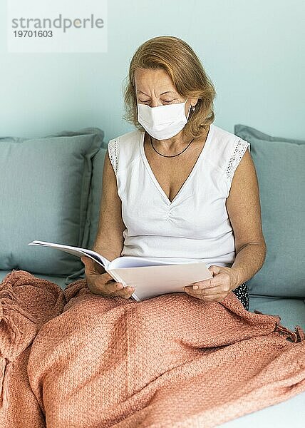 Ältere Frau mit medizinischer Maske  die während der Pandemie zu Hause ein Buch liest