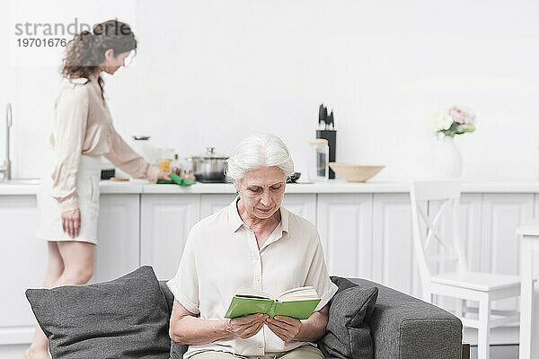 Ältere Frau  die ein Buch liest  vor der Frau  die Hausarbeit verrichtet