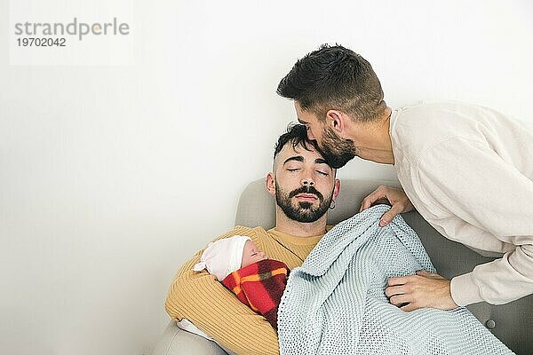 Mann küsst seinen Freund s Stirn schlafen Sofa mit Baby gegen weiße Wand