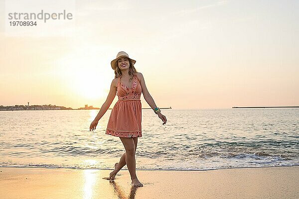 Glückliche Frau in Sommerkleidung  die bei Sonnenuntergang am Rande des Wassers am Strand entlang läuft