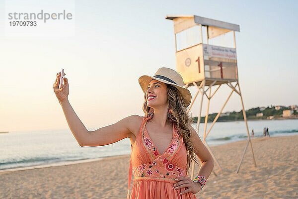 Glückliche Frau mit Sommerkleid und Hut macht ein Selfie am Strand bei Sonnenuntergang