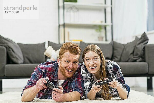 Glückliches junges Paar  das auf einem Teppich liegt und ein Videospiel mit Joystick spielt