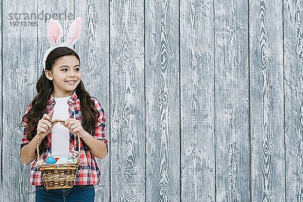 Portrait Mädchen mit Hasenohren hält Ostereierkorb und schaut weg